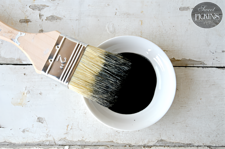 Paintbrush resting on a bowl of Sweet Pickins Black Oil Wax Old Fashioned Milk Paint on a white wooden surface.