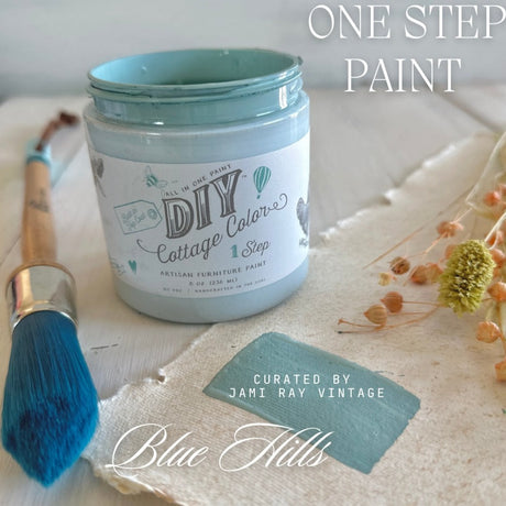 Open jar of Blue Hills DIY Cottage Color by DIY Paint Co. with blue brush and dried flowers on white surface.
