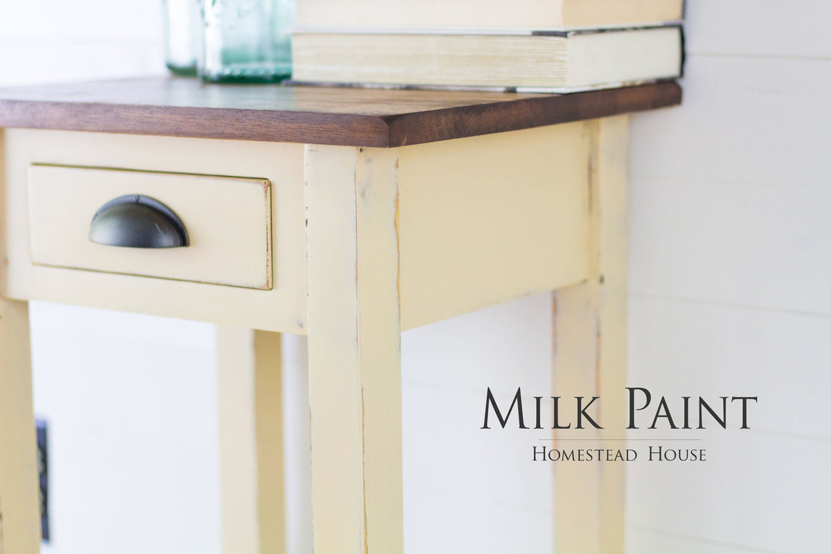 Small table painted in Homestead House Buttermilk Cream with drawer, books, bottles; text reads: Homestead House.