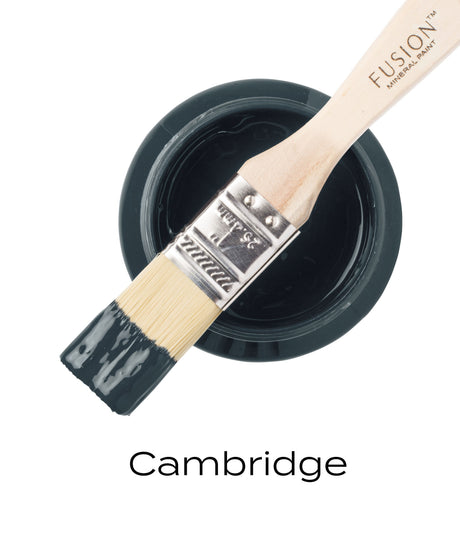 Paintbrush with dark blue paint on open Cambridge Fusion Mineral Paint can, black brand text below.