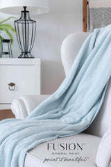 White chair with blue throw, set against Cashmere by Fusion Mineral Paint walls, beside a lamp and plant.