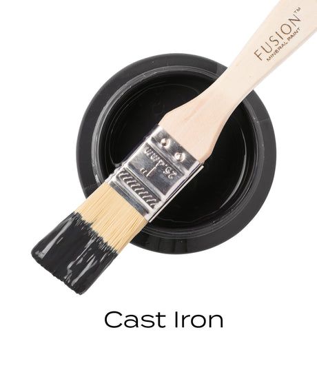 Paintbrush with black paint resting on open jar of Fusion Mineral Paint, label reads “Cast Iron.”.