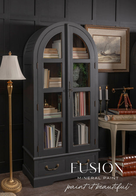 Dark gray arched glass cabinet in Fusion Mineral Paint Cast Iron, filled with books and decor, logo at bottom.