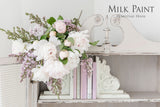 Homestead House Cathedral Taupe Milk Paint can with white and pink flowers and pastel books on a shelf.
