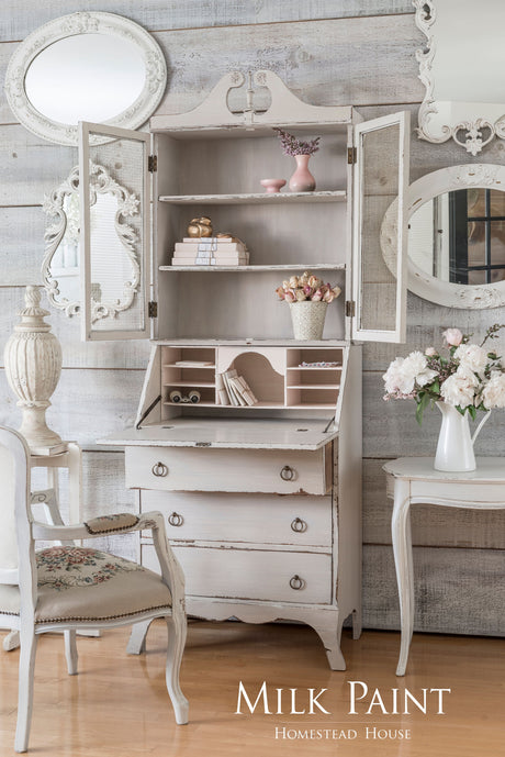 Distressed secretary desk in Cathedral Taupe Milk Paint by Homestead House, shown with white chair and mirrors.