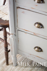 White dresser with metal cup handles, finished in Homestead House Cathedral Taupe Milk Paint, next to a chair.