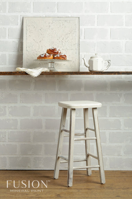 White stool on wood floor beneath shelf with teapot, pastries, and framed panel painted in Champlain Fusion Mineral Paint.