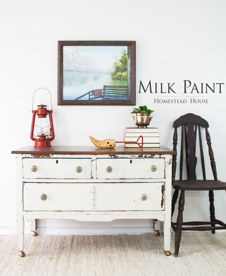 White distressed dresser painted with Champlain Milk Paint by Homestead House, styled with books, lantern, and wall art.