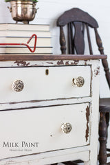 Distressed white dresser in Champlain Milk Paint with floral knobs, stacked books, metal pot, and wooden chair nearby.