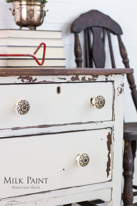 Distressed white dresser in Champlain Milk Paint with floral knobs, stacked books, metal pot, and wooden chair nearby.