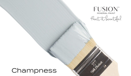 Paintbrush applying Champness Fusion Mineral Paint in light gray-blue shade on white surface.