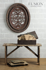 Rustic wooden bench painted Chocolate Fusion Mineral Paint with box on seat; ornate decor hangs above on white brick wall.