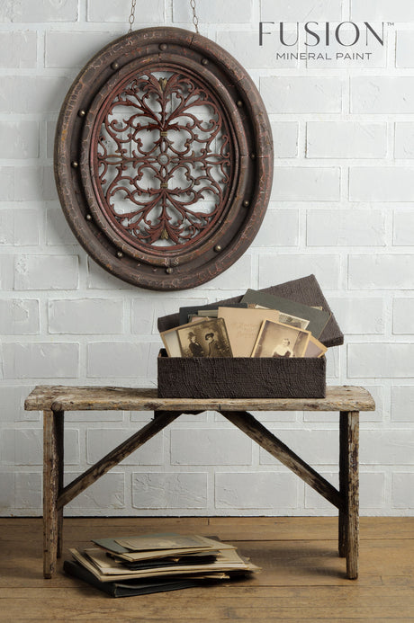 Rustic wooden bench painted Chocolate Fusion Mineral Paint with box on seat; ornate decor hangs above on white brick wall.