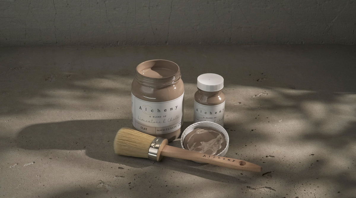 Three jars of Fusion Mineral Paint Clay Alchemy in taupe-gray shades with a brush nearby.