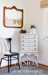 White dresser with gold knobs in Cobblestone Fusion Mineral Paint, shown with mirror, books, gold duck, and chair.