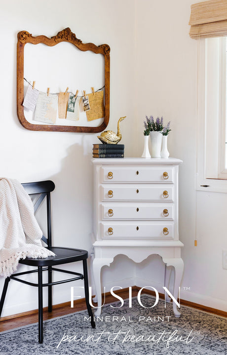 White dresser with gold knobs in Cobblestone Fusion Mineral Paint, shown with mirror, books, gold duck, and chair.