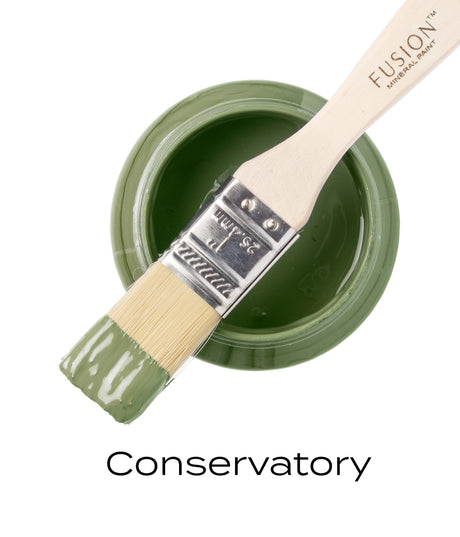 Open jar of Conservatory Fusion Mineral Paint with brush on top, bristles coated in mid-tone green paint.