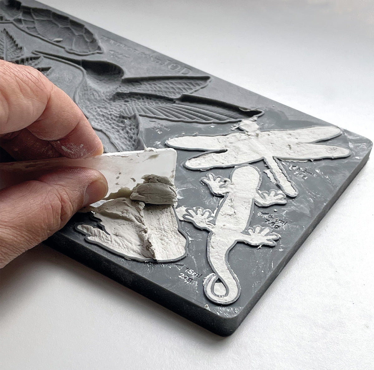 Hand lifting dried plaster from IOD Dewdrop Pond Mould with dragonfly and lizard shapes on a gray surface.