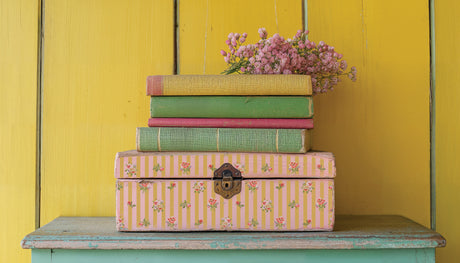 Box containing books and I.O.D. Ditsy Blooms Paint Inlay on a shelf, yellow paneled wall in background.
