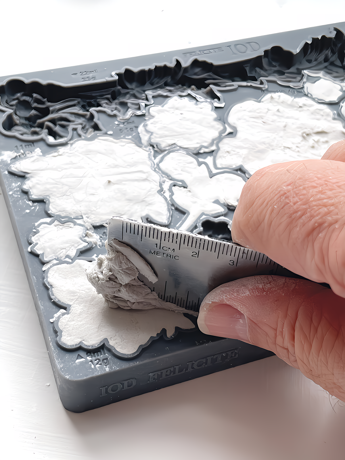 Hand leveling white clay in the Felicite Mould by I.O.D. with a metal ruler on a white surface.