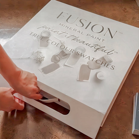 Hands opening Fusion Mineral Paint Briefcase Swatch Box Set labeled “True Colour Swatches” on a brown table.