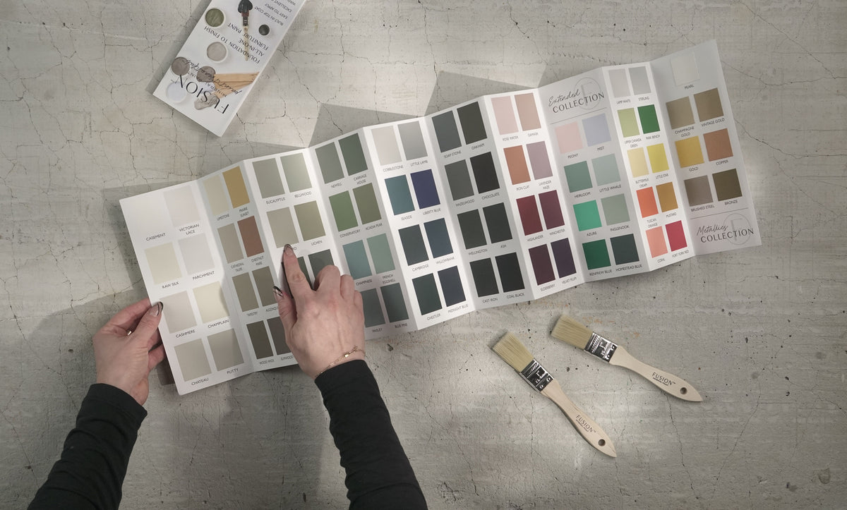 Hand pointing to Fusion Mineral Paint True to Color Card with real paint chips, brushes, and booklets nearby.