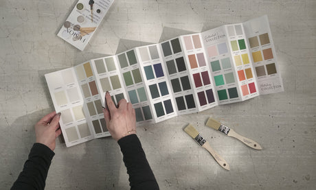 Hand pointing to Fusion Mineral Paint True to Color Card with real paint chips, brushes, and booklets nearby.
