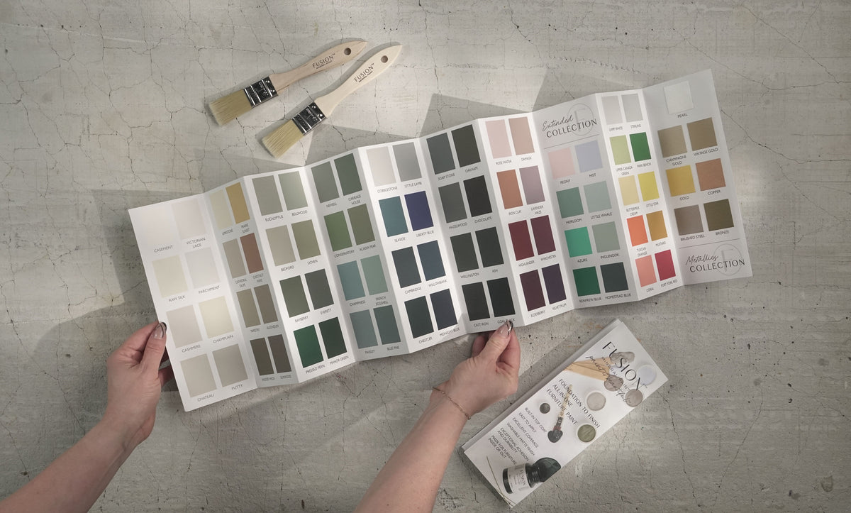 Hands holding a Fusion Mineral Paint True to Color Card with 2024 paint chips and booklet, brushes visible nearby.