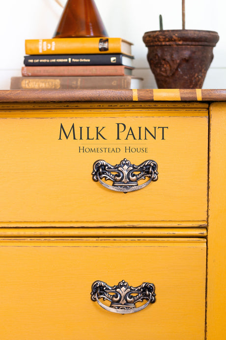Yellow dresser with ornate handles, painted in Homestead House Garden Seed Milk Paint, topped with books and a plant.