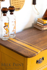 Two glass bottles with orange designs on a yellow Homestead House Garden Seed Milk Paint dresser.