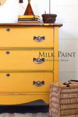 Yellow dresser with ornate handles, books, lamp, plant; wicker basket; text: Garden Seed Milk Paint by Homestead House.