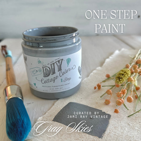 DIY Paint Co. Gray Skies Cottage Color, blue-tipped brush, and dried flowers on a light wood surface.