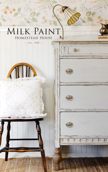 Homestead House Grey Silk Milk Paint on distressed dresser, vintage lamp, lace pillow on chair, floral wallpaper backdrop.