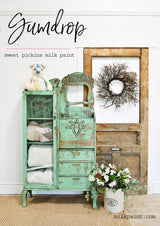 Cabinet painted in Gumdrop by Old Fashioned Milk Paint, storing linens and a teddy, near a door with a wreath.