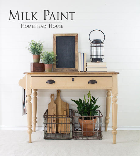 Beige wooden table finished in Homestead House Hampton Milk Paint, shown with plants, books, a lantern, and baskets.