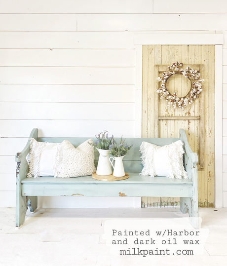 Harbor by Old Fashioned Milk Paint light blue bench with pillows, jug, and lavender near a cream door.