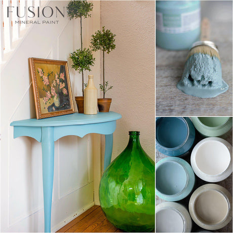 Heirloom Fusion Mineral Paint jars and a blue paintbrush displayed on a table with a green vase.