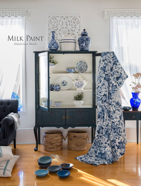 Blue cabinet in Homestead House’s Homestead Blue Milk Paint displaying china, with bowls and floral fabric nearby.