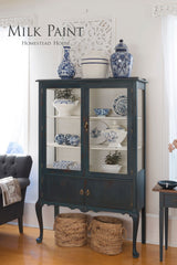 Homestead Blue Milk Paint china cabinet by Homestead House with displayed ceramics and baskets beneath on wood floor.