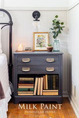 Homestead Grey Milk Paint nightstand with brass pulls, books, artwork, plant, candle, and light above.