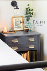 Homestead House Homestead Grey Milk Paint nightstand with lamp, art, books, plant, and candle beside a bed.