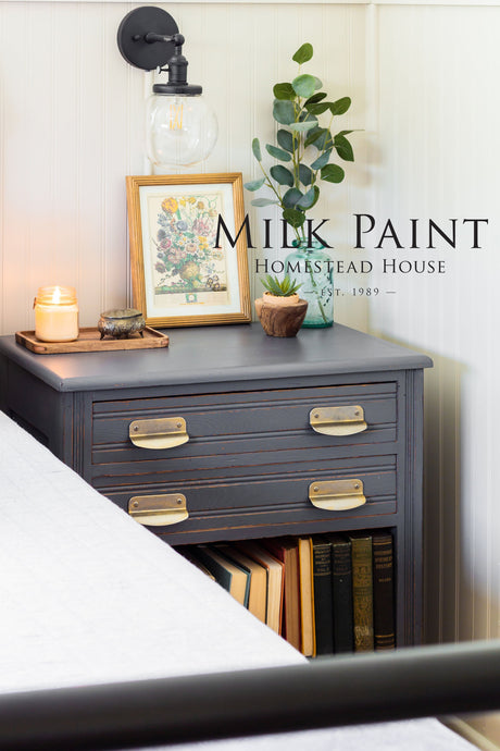 Homestead House Homestead Grey Milk Paint nightstand with lamp, art, books, plant, and candle beside a bed.
