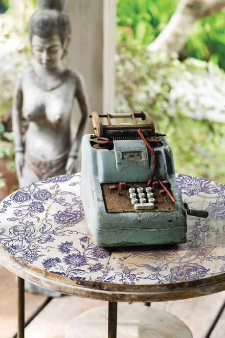 Rusty adding machine on round table featuring I.O.D. Indigo Floral Paint Inlay.