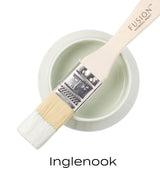 Paintbrush with pale green Inglenook Fusion Mineral Paint resting on a jar labeled Inglenook in black text.