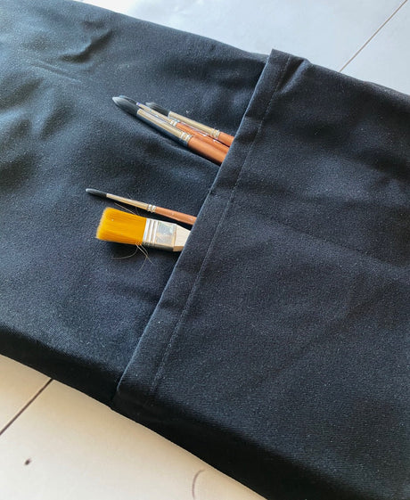 Three paintbrushes in the pocket of an IOD Black Apron by I.O.D., displayed on black fabric.