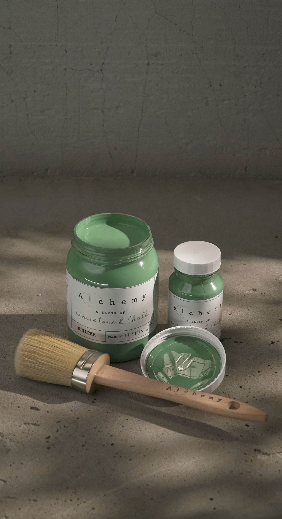Open jar and closed bottle of Juniper Alchemy Paint by Fusion Mineral Paint with brush and lid nearby on gray surface.