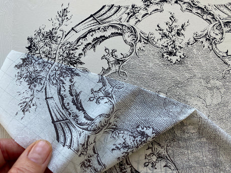 Hand holding fabric with black designs, resembling La Chasse Paint Inlay by IOD shown on decorated furniture below.