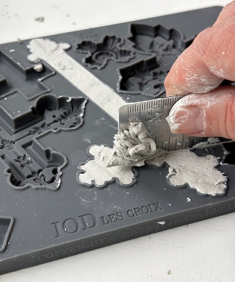 Hand using a ruler to remove clay from Les Croix Mould by IOD on a white surface.