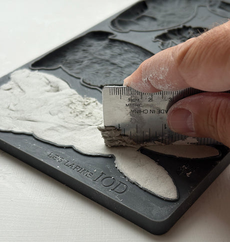 Hand using ruler to level clay in Les Lapins Mould by I.O.D. from Iron Orchid Designs on a white surface.