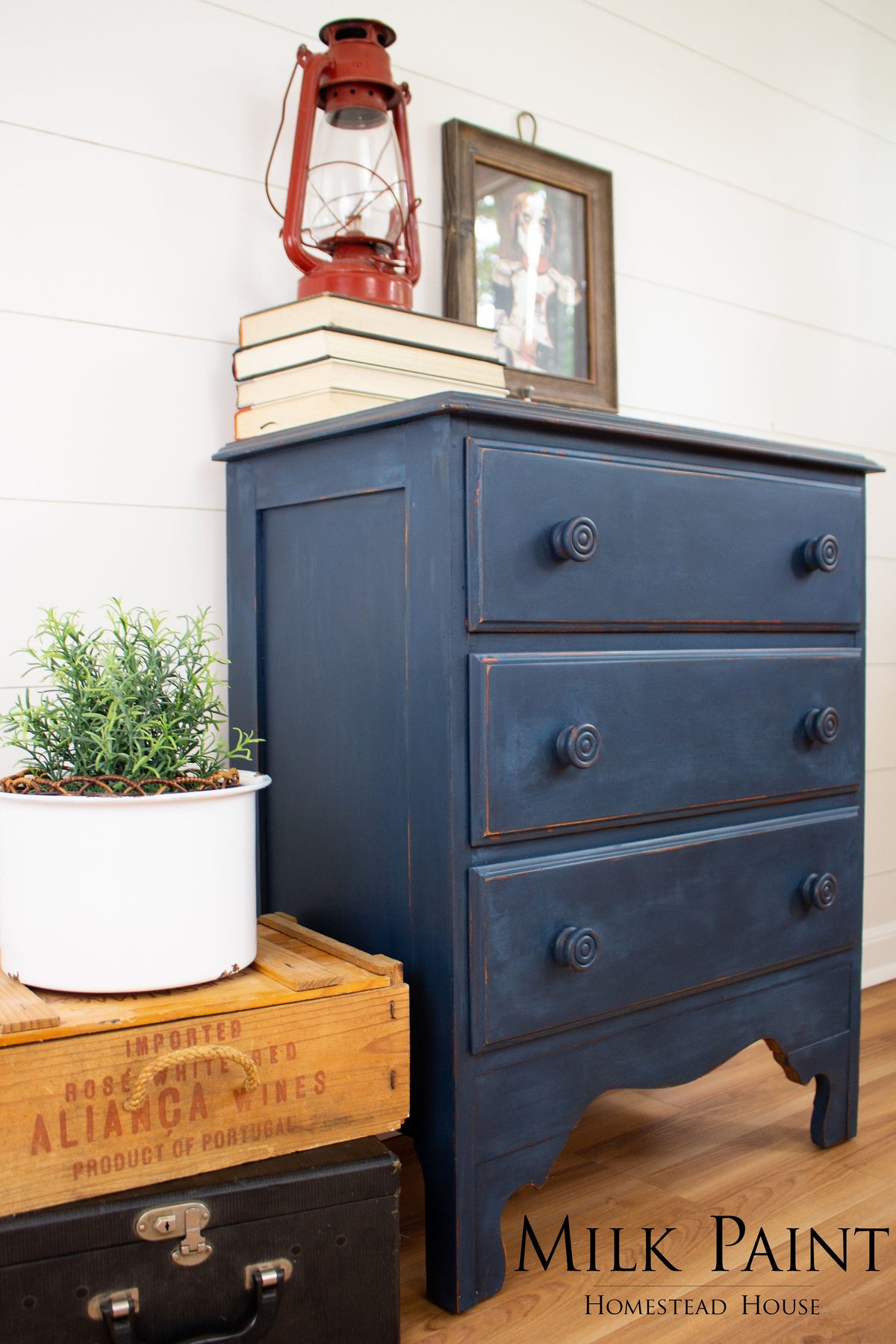 Blue dresser painted with Homestead House Liberty Blue Milk Paint, shown with books, lantern, plant, and boxes nearby.